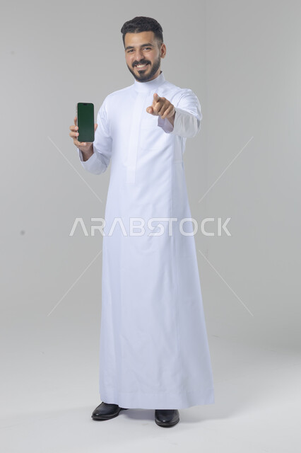  تعابير وايماءات الاندماج والسرور، استخدام التقنية الحديثه المتطورة في الحياة العملية، بورتريه لشاب عربي خليجي سعودي مبتسم يرتدي الثوب التقليدي يقوم بعرض شاشة فارغة على الهاتف المحمول، صورة الجسم بالطول الكامل، خلفية بيضاء