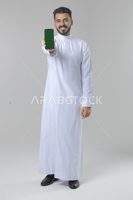  تعابير وايماءات الاندماج والسرور، استخدام التقنية الحديثه المتطورة في الحياة العملية، بورتريه لشاب عربي خليجي سعودي مبتسم يرتدي الثوب التقليدي يقوم بعرض شاشة فارغة على الهاتف المحمول، صورة الجسم بالطول الكامل، خلفية بيضاء
