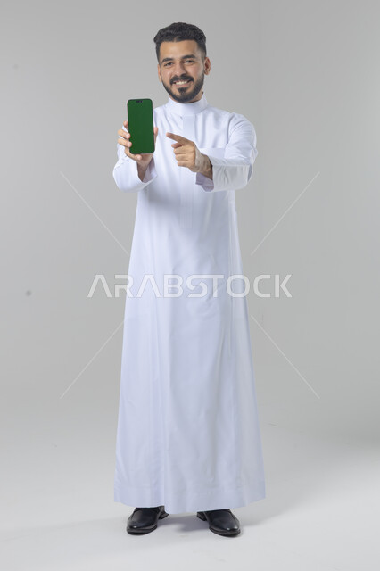  تعابير وايماءات الاندماج والسرور، استخدام التقنية الحديثه المتطورة في الحياة العملية، بورتريه لشاب عربي خليجي سعودي مبتسم يرتدي الثوب التقليدي يقوم بعرض شاشة فارغة على الهاتف المحمول، صورة الجسم بالطول الكامل، خلفية بيضاء