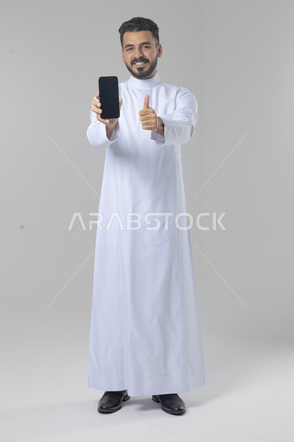  تعابير وايماءات الاندماج والسرور، استخدام التقنية الحديثه المتطورة في الحياة العملية، بورتريه لشاب عربي خليجي سعودي مبتسم يرتدي الثوب التقليدي يقوم بعرض شاشة فارغة على الهاتف المحمول، صورة الجسم بالطول الكامل، خلفية بيضاء