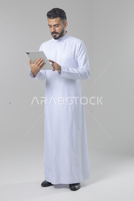 Using modern advanced technology in practical life via tablet, expressions and gestures of integration, portrait of a smiling Saudi Arabian Gulf young man wearing traditional dress working on a tablet, full body image, white background