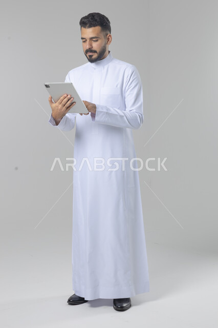استخدام التقنية الحديثه المتطورة في الحياة العملية عبر الجهاز اللوحي، تعابير وايماءات الاندماج، بورتريه لشاب عربي خليجي سعودي مبتسم يرتدي الثوب التقليدي يقوم بالعمل على جهازالتابلت، صورة الجسم بالطول الكامل، خلفية بيضاء