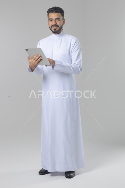 استخدام التقنية الحديثه المتطورة في الحياة العملية عبر الجهاز اللوحي، تعابير وايماءات الاندماج، بورتريه لشاب عربي خليجي سعودي مبتسم يرتدي الثوب التقليدي يقوم بالعمل على جهازالتابلت، صورة الجسم بالطول الكامل، خلفية بيضاء