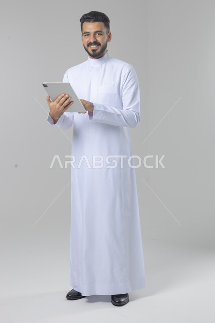 استخدام التقنية الحديثه المتطورة في الحياة العملية عبر الجهاز اللوحي، تعابير وايماءات الاندماج، بورتريه لشاب عربي خليجي سعودي مبتسم يرتدي الثوب التقليدي يقوم بالعمل على جهازالتابلت، صورة الجسم بالطول الكامل، خلفية بيضاء