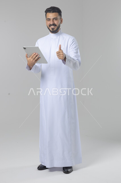 استخدام التقنية الحديثه المتطورة في الحياة العملية عبر الجهاز اللوحي، تعابير وايماءات الاندماج، بورتريه لشاب عربي خليجي سعودي مبتسم يرتدي الثوب التقليدي يقوم بالعمل على جهازالتابلت، صورة الجسم بالطول الكامل، خلفية بيضاء