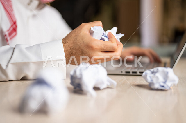 رجل عربي خليجي سعودي يجلس امام الحاسوب المحمول، القيام بتجعيد الأوراق، البحث عن افكار وخطط، الاخفاق في الكتابة والتدوين، الفشل في الابداع والعمل، محاولة ايجاد حلول