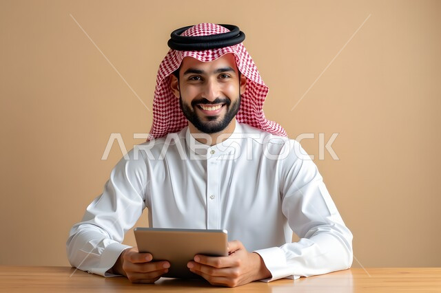 استخدام الأجهزة التكنولوجية الحديثة، بورتريه مقرب لرجل أعمال عربي خليجي سعودي مبتسم يرتدي الشماغ والثوب التقليدي يقوم بالعمل على جهاز التابلت بتعابير السرور، إيماءات تدل على السعادة، مهن ووظائف مكتبية، خلفية بيج