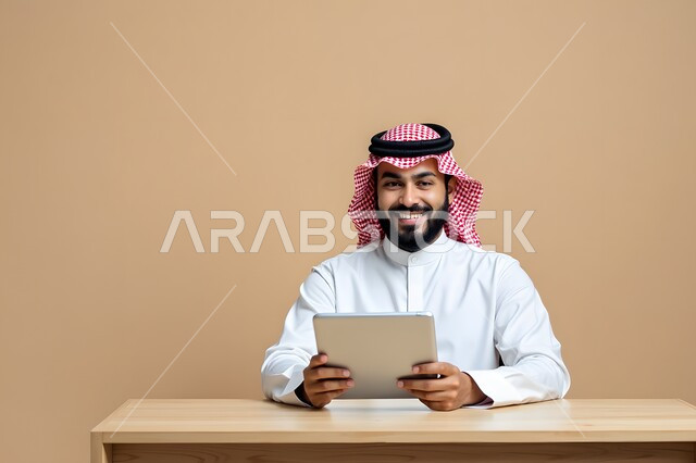 استخدام الأجهزة التكنولوجية الحديثة، بورتريه مقرب لرجل أعمال عربي خليجي سعودي مبتسم يرتدي الشماغ والثوب التقليدي يقوم بالعمل على جهاز التابلت بتعابير السرور، إيماءات تدل على السعادة، مهن ووظائف مكتبية، خلفية بيج