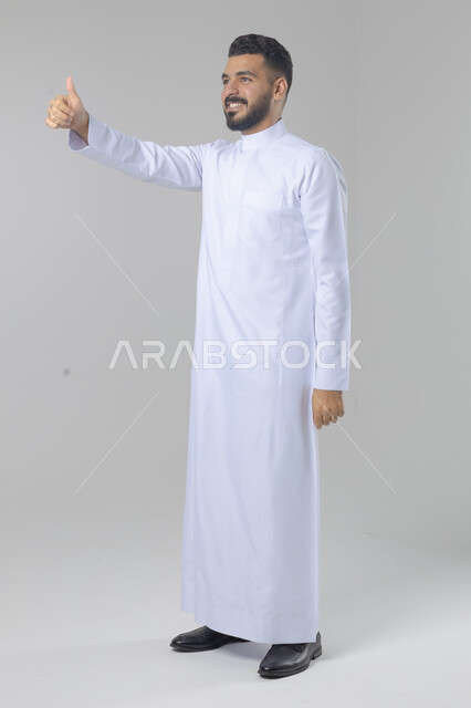  الإمتياز والتميز والنجاح، بورتريه لرجل عربي خليجي سعودي يرتدي الثوب التقليدي ويرفع أصابع الإبهام لأعلى، إيماءات السعادة والإعجاب والموافقة، حركة أصبع تدل على القبول والرضا، الجسم بالطول الكامل، خلفية بيضاء 