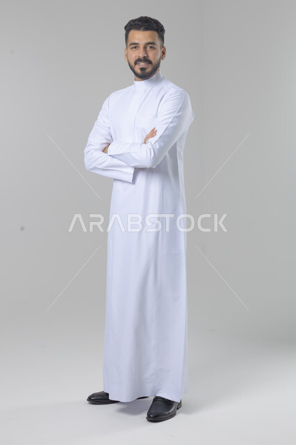  التقاط صور شخصية للوثائق الرسمية، الاعتناء والاهتمام بالمظهر الخارجي، مفهوم الاناقة والرجولة، بورتريه لشاب عربي خليجي سعودي يرتدي الثوب التقليدي يقف بأيدي متقاطعة ينظر الى الكاميرا بتعابير السرور والثقة بالنفس، الجسم بالطول الكامل، خلفية بيضاء