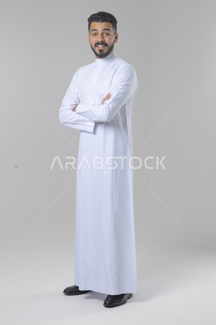  التقاط صور شخصية للوثائق الرسمية، الاعتناء والاهتمام بالمظهر الخارجي، مفهوم الاناقة والرجولة، بورتريه لشاب عربي خليجي سعودي يرتدي الثوب التقليدي يقف بأيدي متقاطعة ينظر الى الكاميرا بتعابير السرور والثقة بالنفس، الجسم بالطول الكامل، خلفية بيضاء