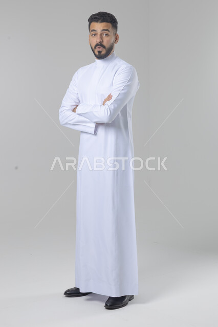  التقاط صور شخصية للوثائق الرسمية، الاعتناء والاهتمام بالمظهر الخارجي، مفهوم الاناقة والرجولة، بورتريه لشاب عربي خليجي سعودي يرتدي الثوب التقليدي يقف بأيدي متقاطعة ينظر الى الكاميرا بتعابير السرور والثقة بالنفس، الجسم بالطول الكامل، خلفية بيضاء