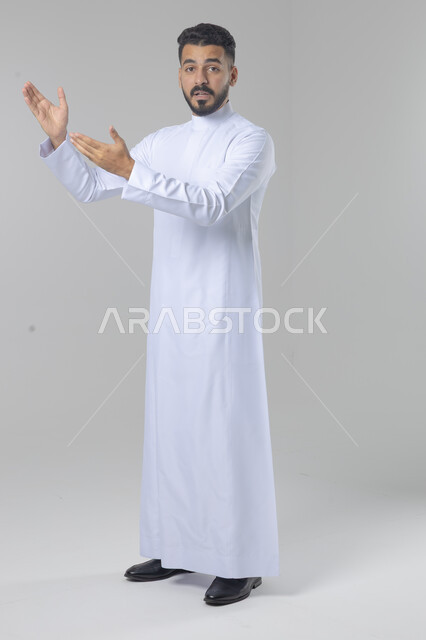 الإشارة إلى شيء ما, الوقوف باستقامة واعتدال, بورتريه لرجل عربي خليجي سعودي يرتدي الثوب التقليدي يشير بيديه لمكان معين, صورة للجسم بالطول الكامل, خلفية بيضاء