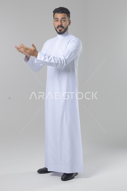 الإشارة إلى شيء ما, الوقوف باستقامة واعتدال, بورتريه لرجل عربي خليجي سعودي يرتدي الثوب التقليدي يشير بيديه لمكان معين, صورة للجسم بالطول الكامل, خلفية بيضاء