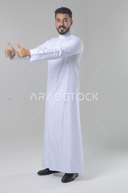  الإمتياز والتميز والنجاح، بورتريه لرجل عربي خليجي سعودي يرتدي الثوب التقليدي ويرفع أصابع الإبهام لأعلى، إيماءات السعادة والإعجاب والموافقة، حركة أصبع تدل على القبول والرضا، الجسم بالطول الكامل، خلفية بيضاء 