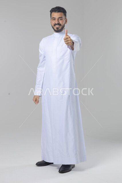  الإمتياز والتميز والنجاح، بورتريه لرجل عربي خليجي سعودي يرتدي الثوب التقليدي ويرفع أصابع الإبهام لأعلى، إيماءات السعادة والإعجاب والموافقة، حركة أصبع تدل على القبول والرضا، الجسم بالطول الكامل، خلفية بيضاء 