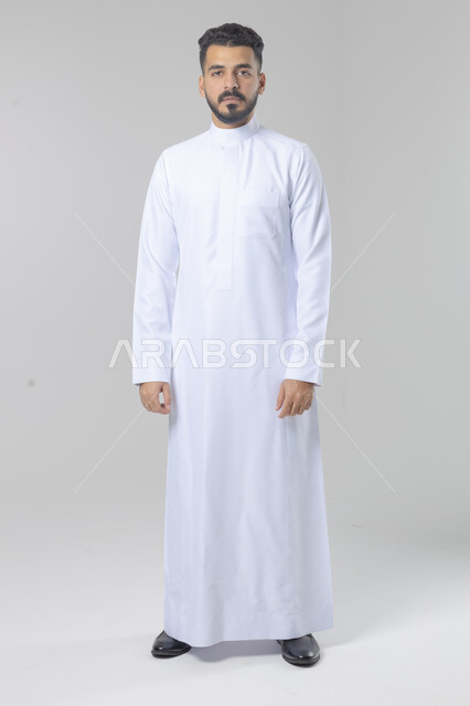 الاعتناء والاهتمام بالمظهر الخارجي، مفهوم الاناقة والرجولة، النظر الى شيء ما، بورتريه لشاب عربي خليجي سعودي يرتدي الثوب التقليدي يقف باستقامة بتعابير السرور والثقة بالنفس، الجسم بالطول الكامل، خلفية بيضاء