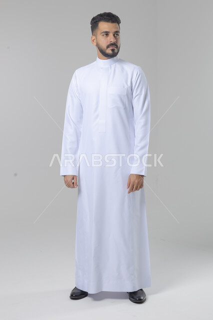 الاعتناء والاهتمام بالمظهر الخارجي، مفهوم الاناقة والرجولة، النظر الى شيء ما، بورتريه لشاب عربي خليجي سعودي يرتدي الثوب التقليدي يقف باستقامة بتعابير السرور والثقة بالنفس، الجسم بالطول الكامل، خلفية بيضاء