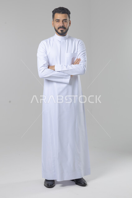  التقاط صور شخصية للوثائق الرسمية، الاعتناء والاهتمام بالمظهر الخارجي، مفهوم الاناقة والرجولة، بورتريه لشاب عربي خليجي سعودي يرتدي الثوب التقليدي يقف بأيدي متقاطعة ينظر الى الكاميرا بتعابير السرور والثقة بالنفس، الجسم بالطول الكامل، خلفية بيضاء