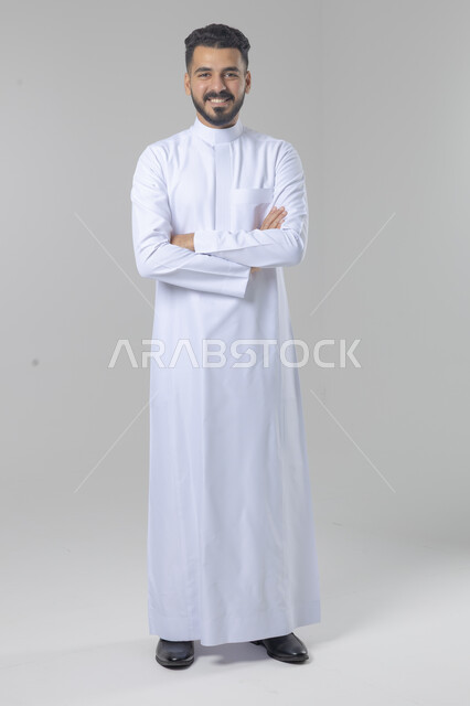  التقاط صور شخصية للوثائق الرسمية، الاعتناء والاهتمام بالمظهر الخارجي، مفهوم الاناقة والرجولة، بورتريه لشاب عربي خليجي سعودي يرتدي الثوب التقليدي يقف بأيدي متقاطعة ينظر الى الكاميرا بتعابير السرور والثقة بالنفس، الجسم بالطول الكامل، خلفية بيضاء