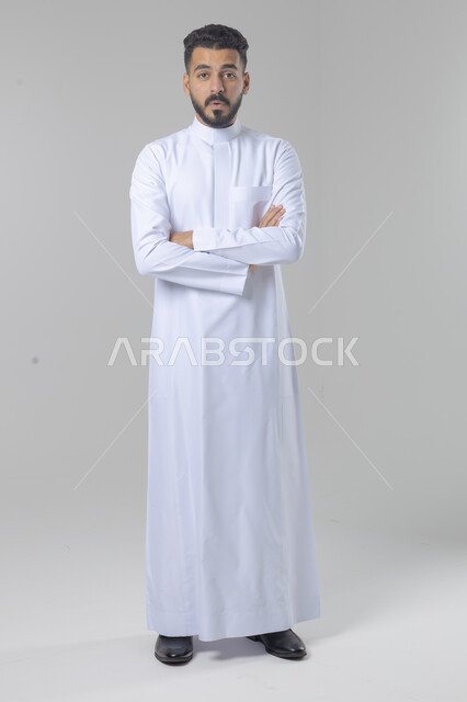  التقاط صور شخصية للوثائق الرسمية، الاعتناء والاهتمام بالمظهر الخارجي، مفهوم الاناقة والرجولة، بورتريه لشاب عربي خليجي سعودي يرتدي الثوب التقليدي يقف بأيدي متقاطعة ينظر الى الكاميرا بتعابير السرور والثقة بالنفس، الجسم بالطول الكامل، خلفية بيضاء