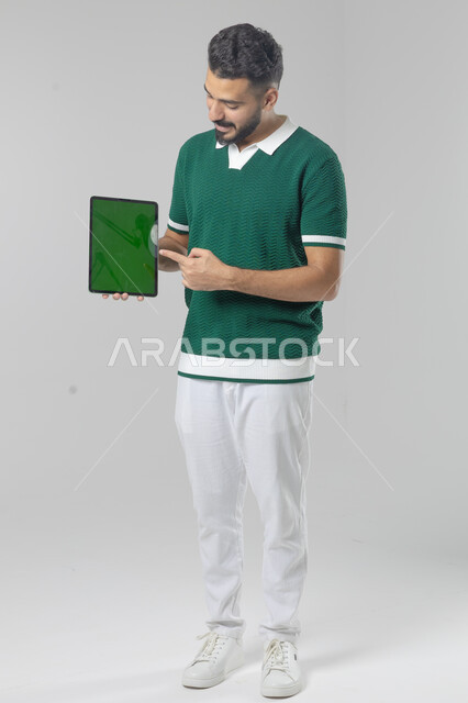 عرض شاشة خضراء كروما عبر الجهاز اللوحي، استعمال التقنية الحديثه المتطورة في الحياة العملية، تعابير وايماءات الاندماج، بورتريه لشاب عربي خليجي سعودي مبتسم يرتدي الزي الكاجوال يقوم بالعمل على جهازالتابلت، صورة الجسم بالطول الكامل، خلفية بيضاء