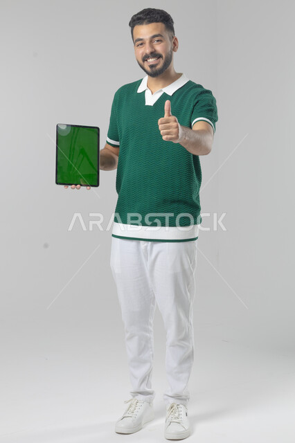 عرض شاشة خضراء كروما عبر الجهاز اللوحي، استعمال التقنية الحديثه المتطورة في الحياة العملية، تعابير وايماءات الاندماج، بورتريه لشاب عربي خليجي سعودي مبتسم يرتدي الزي الكاجوال يقوم بالعمل على جهازالتابلت، صورة الجسم بالطول الكامل، خلفية بيضاء