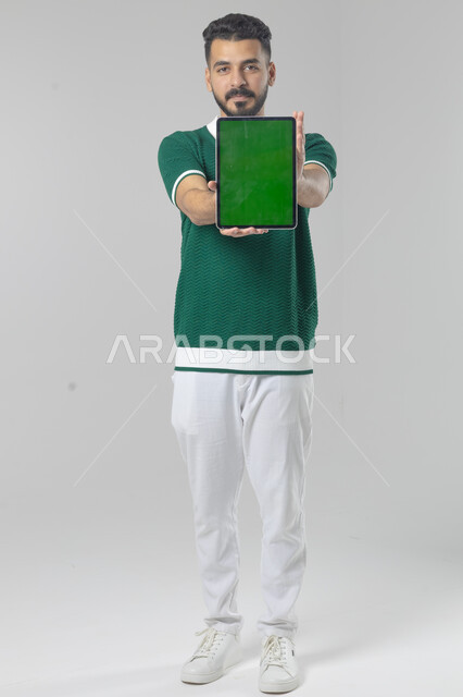 عرض شاشة خضراء كروما عبر الجهاز اللوحي، استعمال التقنية الحديثه المتطورة في الحياة العملية، تعابير وايماءات الاندماج، بورتريه لشاب عربي خليجي سعودي مبتسم يرتدي الزي الكاجوال يقوم بالعمل على جهازالتابلت، صورة الجسم بالطول الكامل، خلفية بيضاء