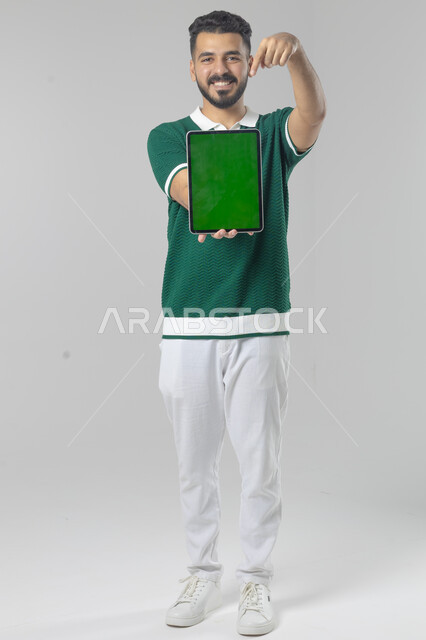 عرض شاشة خضراء كروما عبر الجهاز اللوحي، استعمال التقنية الحديثه المتطورة في الحياة العملية، تعابير وايماءات الاندماج، بورتريه لشاب عربي خليجي سعودي مبتسم يرتدي الزي الكاجوال يقوم بالعمل على جهازالتابلت، صورة الجسم بالطول الكامل، خلفية بيضاء