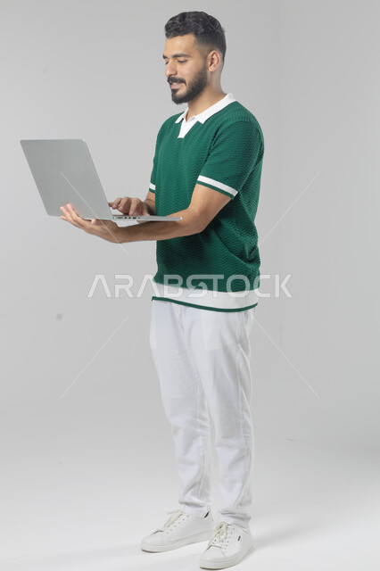 استخدام التقنية الحديثه المتطورة في الحياة العملية، تعابير وايماءات الاندماج والسرور، بورتريه لشاب عربي خليجي سعودي مبتسم يرتدي الزي الكاجوال يقوم بالعمل على الحاسب المحمول، صورة الجسم بالطول الكامل، خلفية بيضاء