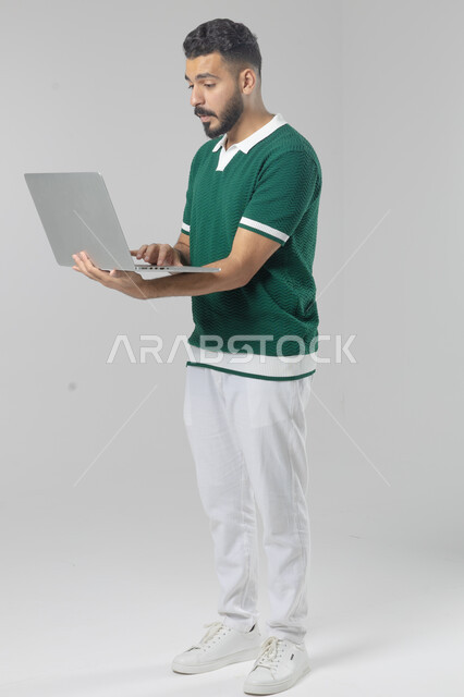Using modern advanced technology in practical life, expressions and gestures of integration and pleasure, portrait of a smiling young Saudi Arabian Gulf man wearing casual clothes working on a laptop, full body image, white background