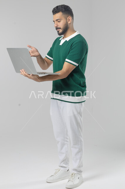 Using modern advanced technology in practical life, expressions and gestures of integration and pleasure, portrait of a smiling young Saudi Arabian Gulf man wearing casual clothes working on a laptop, full body image, white background