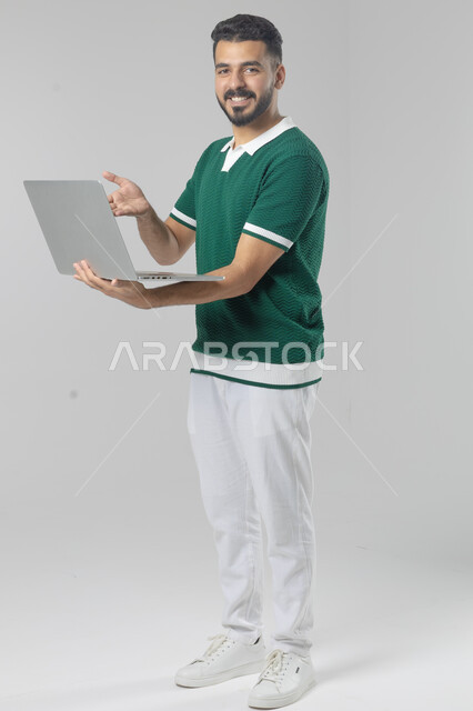 Using modern advanced technology in practical life, expressions and gestures of integration and pleasure, portrait of a smiling young Saudi Arabian Gulf man wearing casual clothes working on a laptop, full body image, white background
