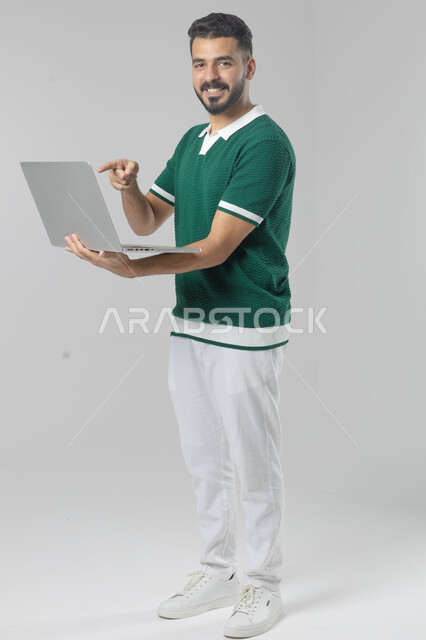 Using modern advanced technology in practical life, expressions and gestures of integration and pleasure, portrait of a smiling young Saudi Arabian Gulf man wearing casual clothes working on a laptop, full body image, white background