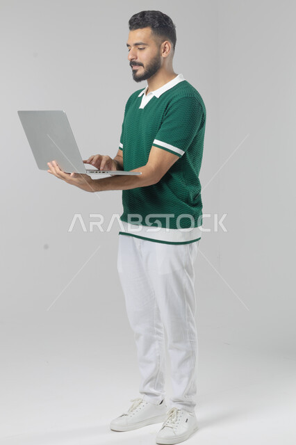 Using modern advanced technology in practical life, expressions and gestures of integration and pleasure, portrait of a smiling young Saudi Arabian Gulf man wearing casual clothes working on a laptop, full body image, white background