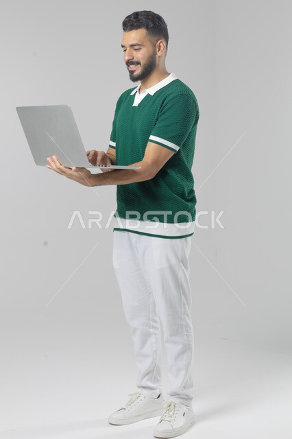 استخدام التقنية الحديثه المتطورة في الحياة العملية، تعابير وايماءات الاندماج والسرور، بورتريه لشاب عربي خليجي سعودي مبتسم يرتدي الزي الكاجوال يقوم بالعمل على الحاسب المحمول، صورة الجسم بالطول الكامل، خلفية بيضاء
