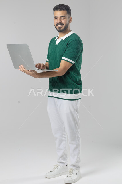 استخدام التقنية الحديثه المتطورة في الحياة العملية، تعابير وايماءات الاندماج والسرور، بورتريه لشاب عربي خليجي سعودي مبتسم يرتدي الزي الكاجوال يقوم بالعمل على الحاسب المحمول، صورة الجسم بالطول الكامل، خلفية بيضاء
