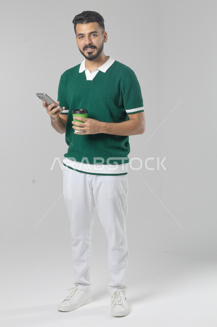 تناول وشرب المشروبات الساخنة، استخدام التقنية الحديثه المتطورة في الحياة العملية، بورتريه لشاب عربي خليجي سعودي مبتسم يرتدي الزي الكاجوال يقوم بالعمل على الهاتف المحمول، صورة الجسم بالطول الكامل، خلفية بيضاء