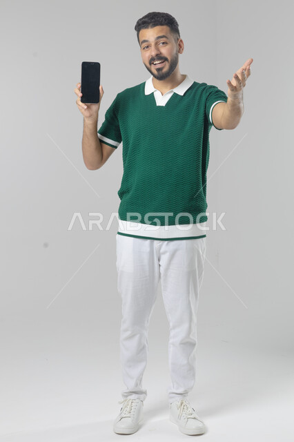  تعابير وايماءات الاندماج والسرور، استخدام التقنية الحديثه المتطورة في الحياة العملية، بورتريه لشاب عربي خليجي سعودي مبتسم يرتدي الزي الكاجوال يقوم بالعمل على الهاتف المحمول، صورة الجسم بالطول الكامل، خلفية بيضاء