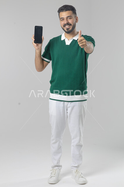  تعابير وايماءات الاندماج والسرور، استخدام التقنية الحديثه المتطورة في الحياة العملية، بورتريه لشاب عربي خليجي سعودي مبتسم يرتدي الزي الكاجوال يقوم بالعمل على الهاتف المحمول، صورة الجسم بالطول الكامل، خلفية بيضاء