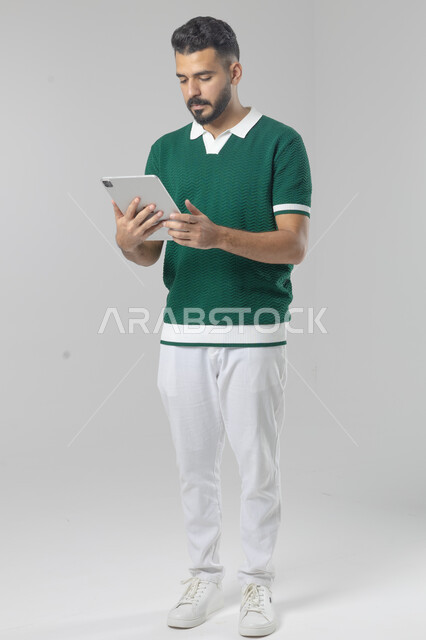 استعمال التقنية الحديثه المتطورة في الحياة العملية عبر الجهاز اللوحي، تعابير وايماءات الاندماج، بورتريه لشاب عربي خليجي سعودي مبتسم يرتدي الزي الكاجوال يقوم بالعمل على جهازالتابلت، صورة الجسم بالطول الكامل، خلفية بيضاء