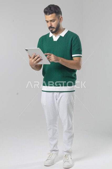 استعمال التقنية الحديثه المتطورة في الحياة العملية عبر الجهاز اللوحي، تعابير وايماءات الاندماج، بورتريه لشاب عربي خليجي سعودي مبتسم يرتدي الزي الكاجوال يقوم بالعمل على جهازالتابلت، صورة الجسم بالطول الكامل، خلفية بيضاء