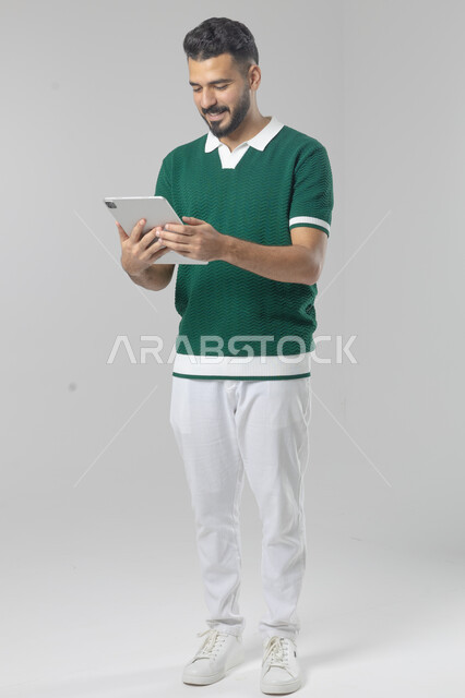 استعمال التقنية الحديثه المتطورة في الحياة العملية عبر الجهاز اللوحي، تعابير وايماءات الاندماج، بورتريه لشاب عربي خليجي سعودي مبتسم يرتدي الزي الكاجوال يقوم بالعمل على جهازالتابلت، صورة الجسم بالطول الكامل، خلفية بيضاء