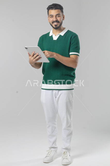 استعمال التقنية الحديثه المتطورة في الحياة العملية عبر الجهاز اللوحي، تعابير وايماءات الاندماج، بورتريه لشاب عربي خليجي سعودي مبتسم يرتدي الزي الكاجوال يقوم بالعمل على جهازالتابلت، صورة الجسم بالطول الكامل، خلفية بيضاء