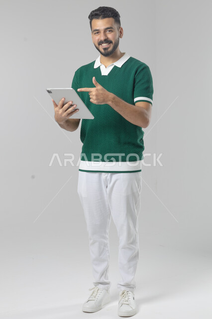استعمال التقنية الحديثه المتطورة في الحياة العملية عبر الجهاز اللوحي، تعابير وايماءات الاندماج، بورتريه لشاب عربي خليجي سعودي مبتسم يرتدي الزي الكاجوال يقوم بالعمل على جهازالتابلت، صورة الجسم بالطول الكامل، خلفية بيضاء