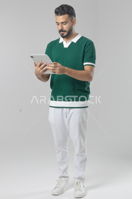 استعمال التقنية الحديثه المتطورة في الحياة العملية عبر الجهاز اللوحي، تعابير وايماءات الاندماج، بورتريه لشاب عربي خليجي سعودي مبتسم يرتدي الزي الكاجوال يقوم بالعمل على جهازالتابلت، صورة الجسم بالطول الكامل، خلفية بيضاء