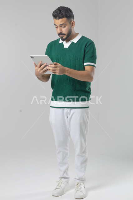 استعمال التقنية الحديثه المتطورة في الحياة العملية عبر الجهاز اللوحي، تعابير وايماءات الاندماج، بورتريه لشاب عربي خليجي سعودي مبتسم يرتدي الزي الكاجوال يقوم بالعمل على جهازالتابلت، صورة الجسم بالطول الكامل، خلفية بيضاء