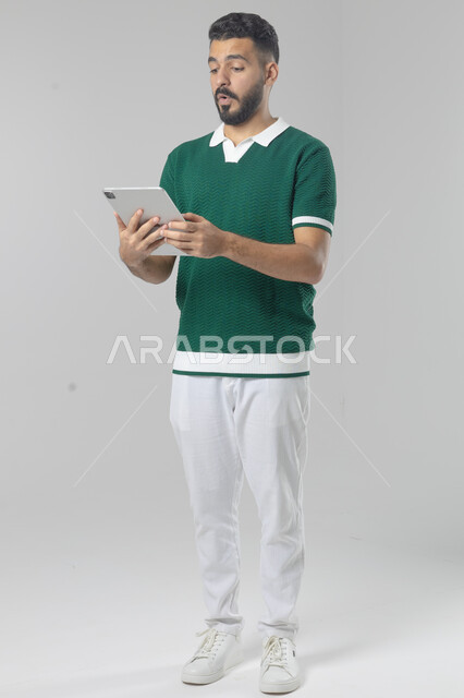 Using modern advanced technology in practical life via tablet, expressions and gestures of integration, portrait of a smiling young Saudi Arabian Gulf man wearing a Ka uniform working on a tablet, full body image, white background