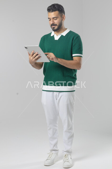 استعمال التقنية الحديثه المتطورة في الحياة العملية عبر الجهاز اللوحي، تعابير وايماءات الاندماج، بورتريه لشاب عربي خليجي سعودي مبتسم يرتدي الزي الكاجوال يقوم بالعمل على جهازالتابلت، صورة الجسم بالطول الكامل، خلفية بيضاء