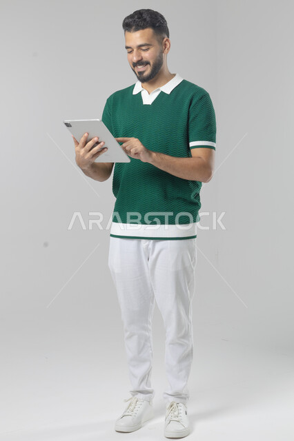 استعمال التقنية الحديثه المتطورة في الحياة العملية عبر الجهاز اللوحي، تعابير وايماءات الاندماج، بورتريه لشاب عربي خليجي سعودي مبتسم يرتدي الزي الكاجوال يقوم بالعمل على جهازالتابلت، صورة الجسم بالطول الكامل، خلفية بيضاء